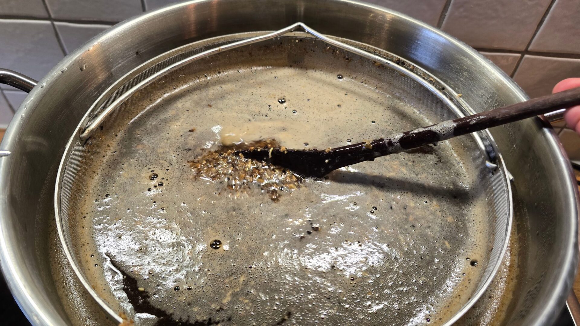 Dunkle Bierwürze mit Schaum und Treber in einem Sieb über einem Edelstahltopf, ein Holzlöffel schiebt den Treber zusammen