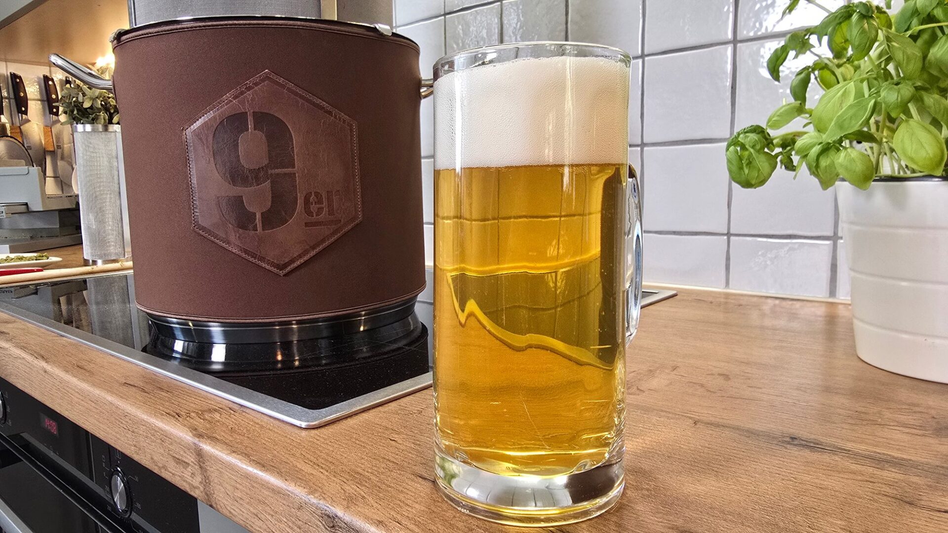 Glaskrug mit hellem Bier und Schaum vor dem 9er Braukessel mit Isoliermantel auf dem Induktionsfeld in der Küche