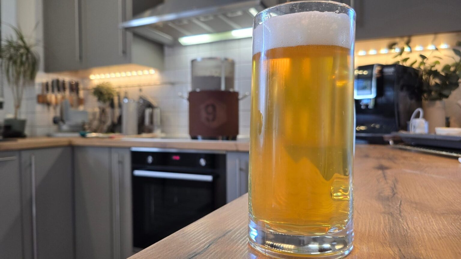 Hohes Glas mit klarem goldenem Bier und Schaumkrone auf Holzplatte, dahinter der 9er Braukessel in einer Küche