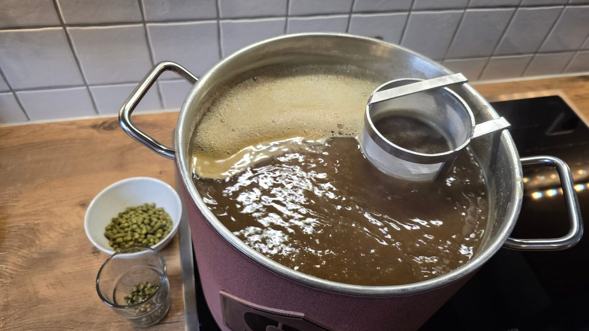 Kochende Würze mit Schaum im Kessel beim Bierbrauen auf dem Herd, daneben Hopfenpellets in Schale und Glas sowie ein Sieb am Rand