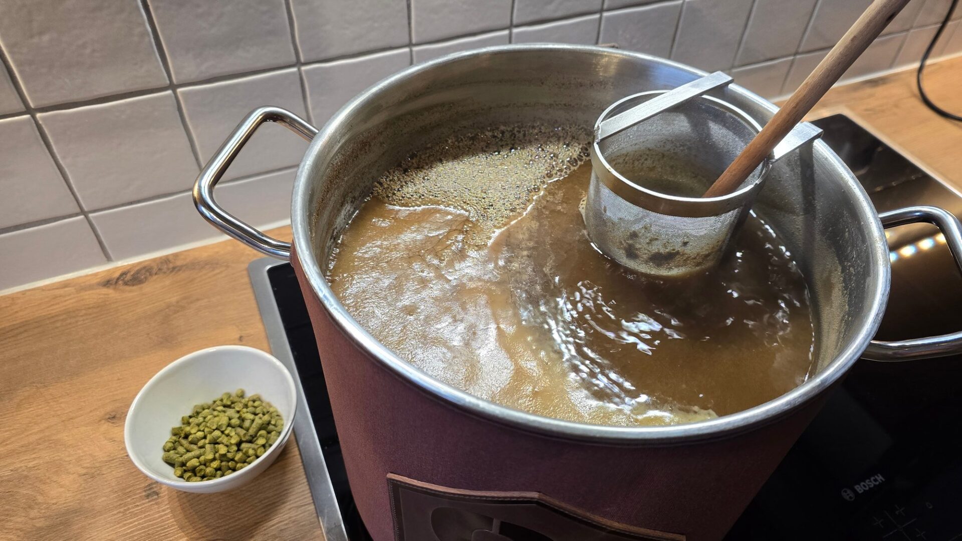 Kochende Würze im Topf auf dem Induktionsherd in der Küche mit Sieblöffel und einer Schale Hopfenpellets daneben