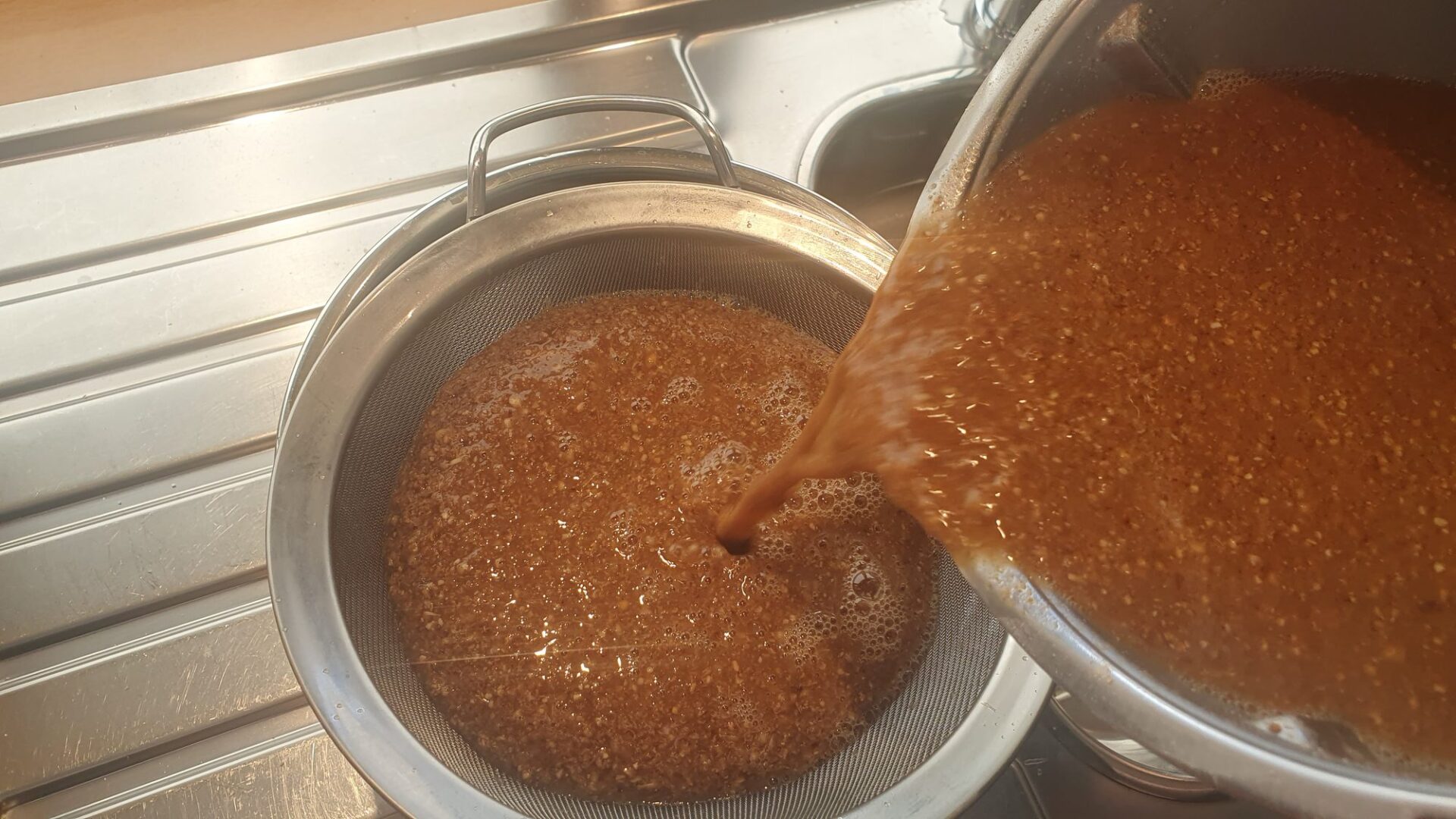 Maische wird aus einem Edelstahltopf durch ein Sieb über einer Küchenspüle gegossen