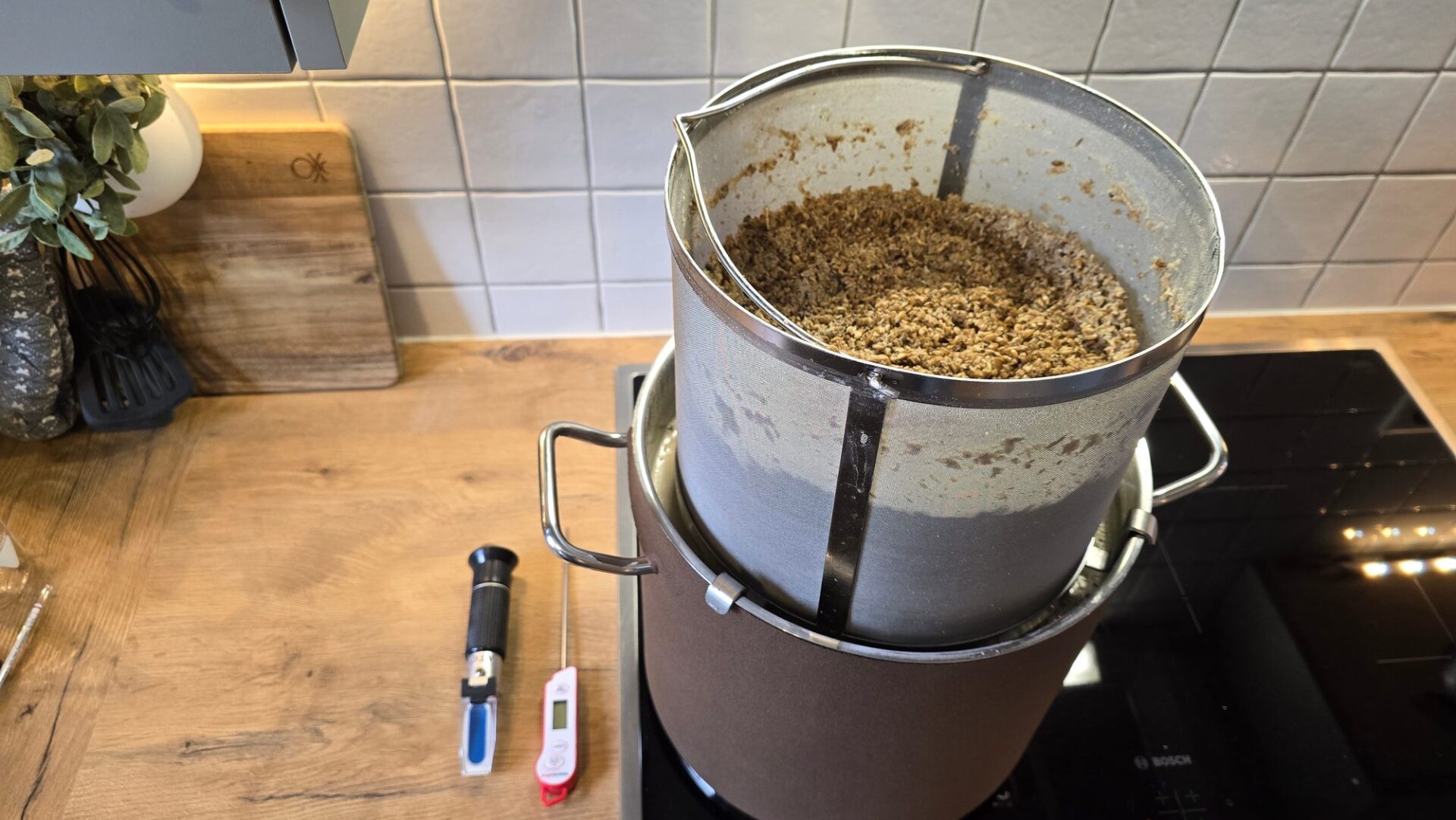Malzrohr mit Treber auf dem 9er Braukessel am Herd in der Küche, daneben Refraktometer und Thermometer auf der Arbeitsplatte