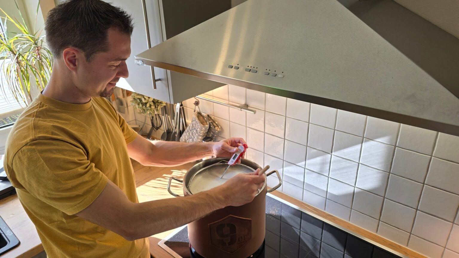 Mann misst mit einem Thermometer die Maische im 9er Braukessel auf dem Induktionsherd in der Küche