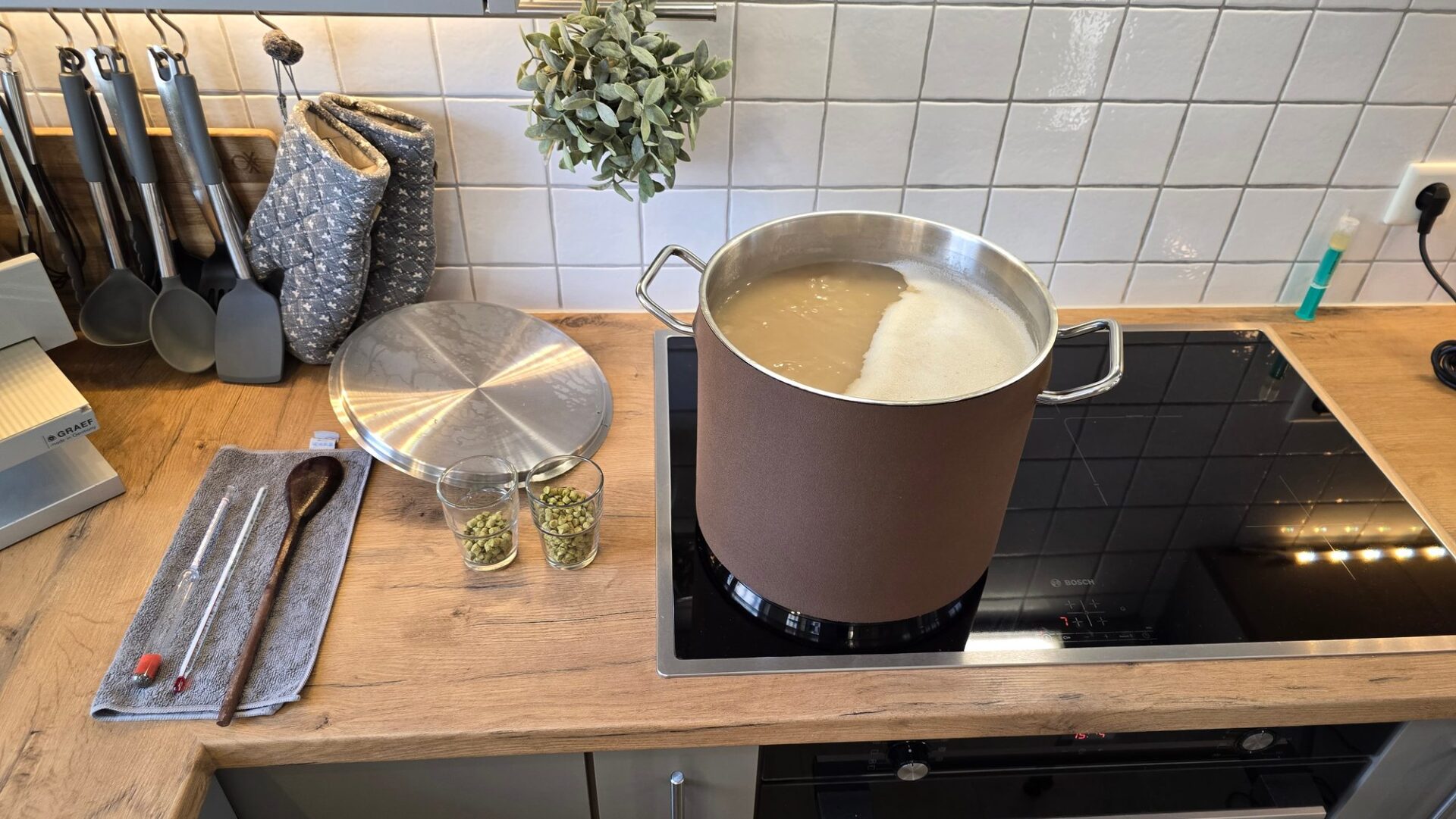 Topf mit kochender Würze auf dem Induktionsfeld, daneben zwei Gläser Hopfenpellets, Holzlöffel und Spindeln auf einem Tuch