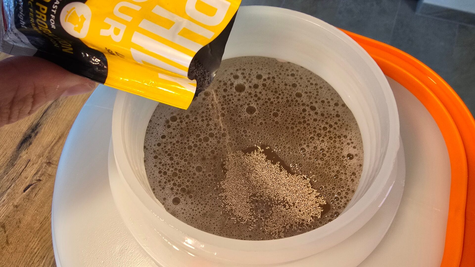 Trockenhefe rieselt aus einem gelben Päckchen auf die schäumende Bierwürze in einem weißen Gäreimer