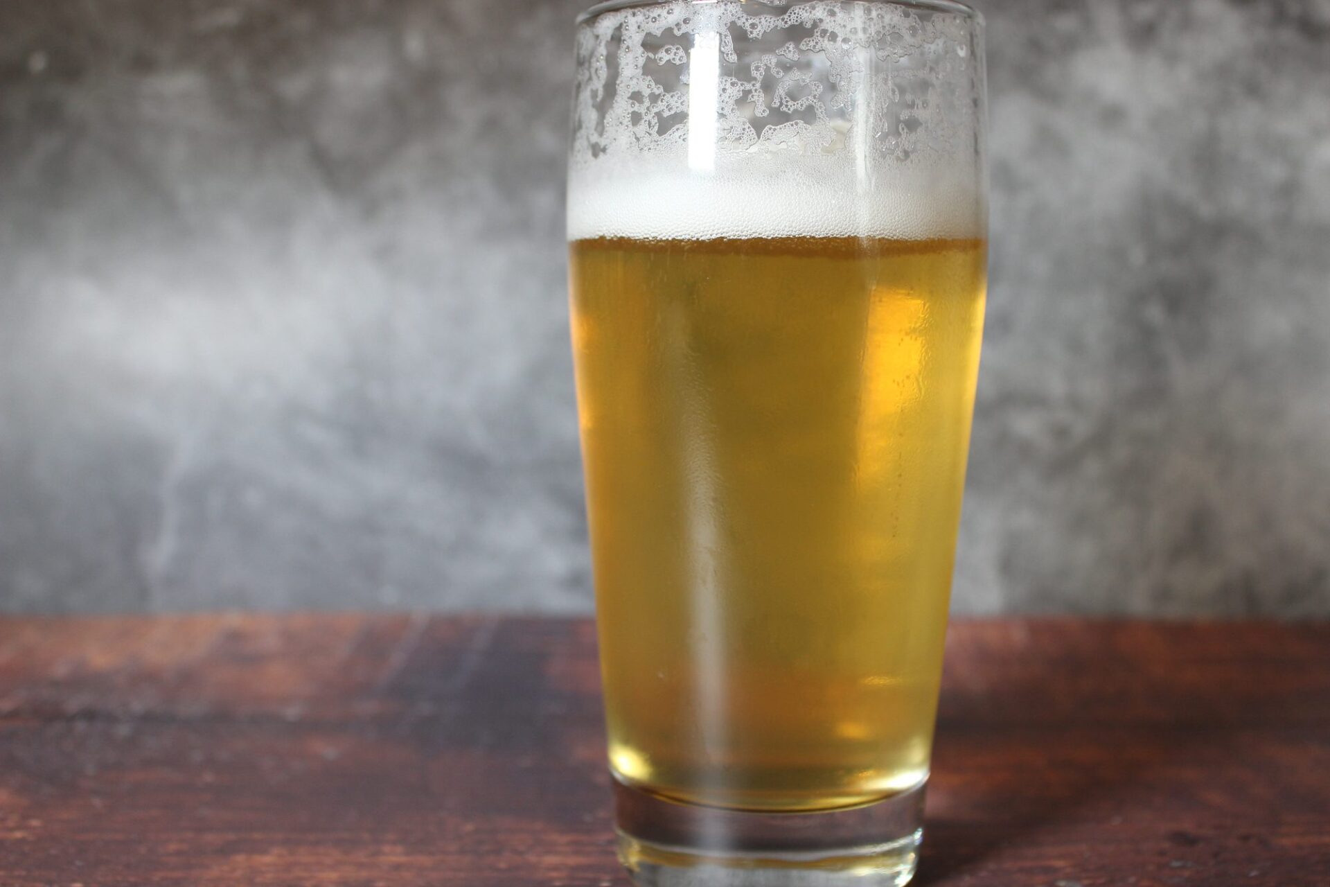 Willybecher mit klarem goldgelbem Bier und Schaumkrone auf dunklem Holztisch vor grauer Betonwand